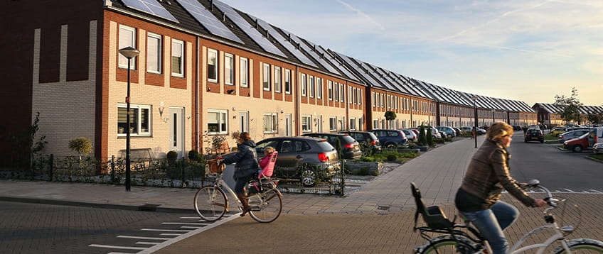 Straat rijtjeshuizen met zonnepanelen. Voor de huizen staan auto's op de oprit. er fietsen twee fietsers in beeld