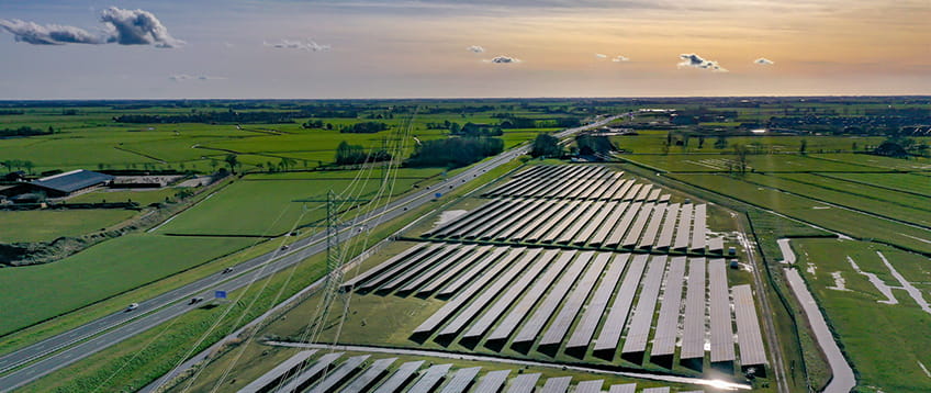 Groot veld zonnepanelen omringd door weilanden. Aan de linkerkant een provinciale autoweg en hoogpanningslijnen