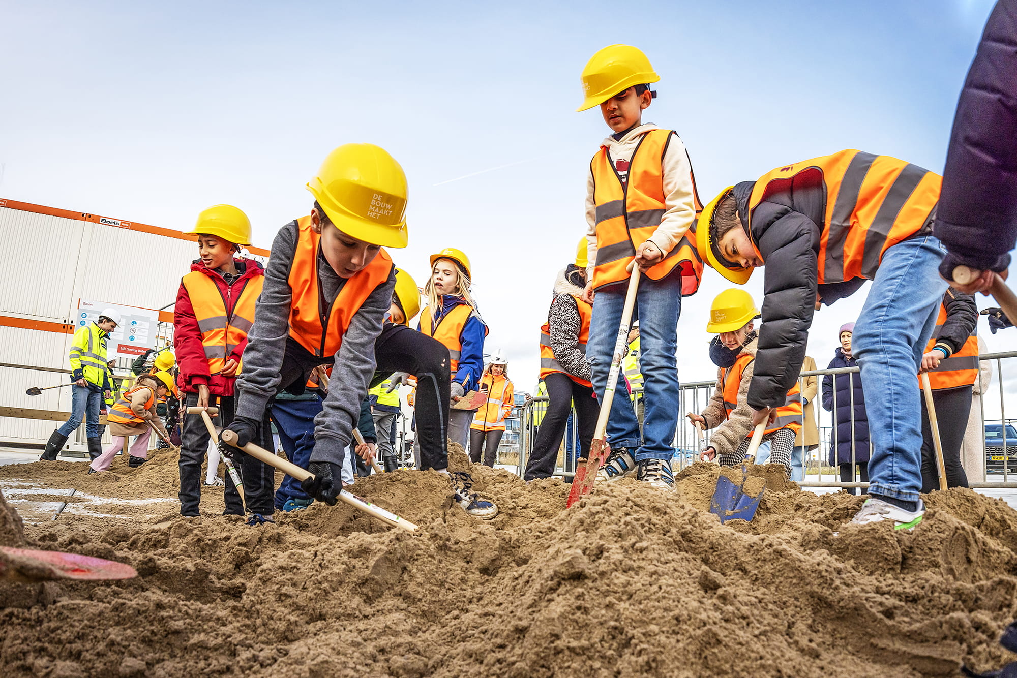 Kinderen staan met een schop bij een berg zand. Ze dragen een veiligheidshesje en helm. Ze zijn enthousiast aan het scheppen.